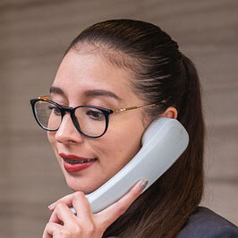 gros plan d'une femme aux cheveux foncés tirés en arrière, portant des lunettes et un haut foncé. Elle tient un récepteur de téléphone de couleur claire à son oreille