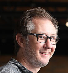 A man with greying hair and glasses, smiling and looking to his left.